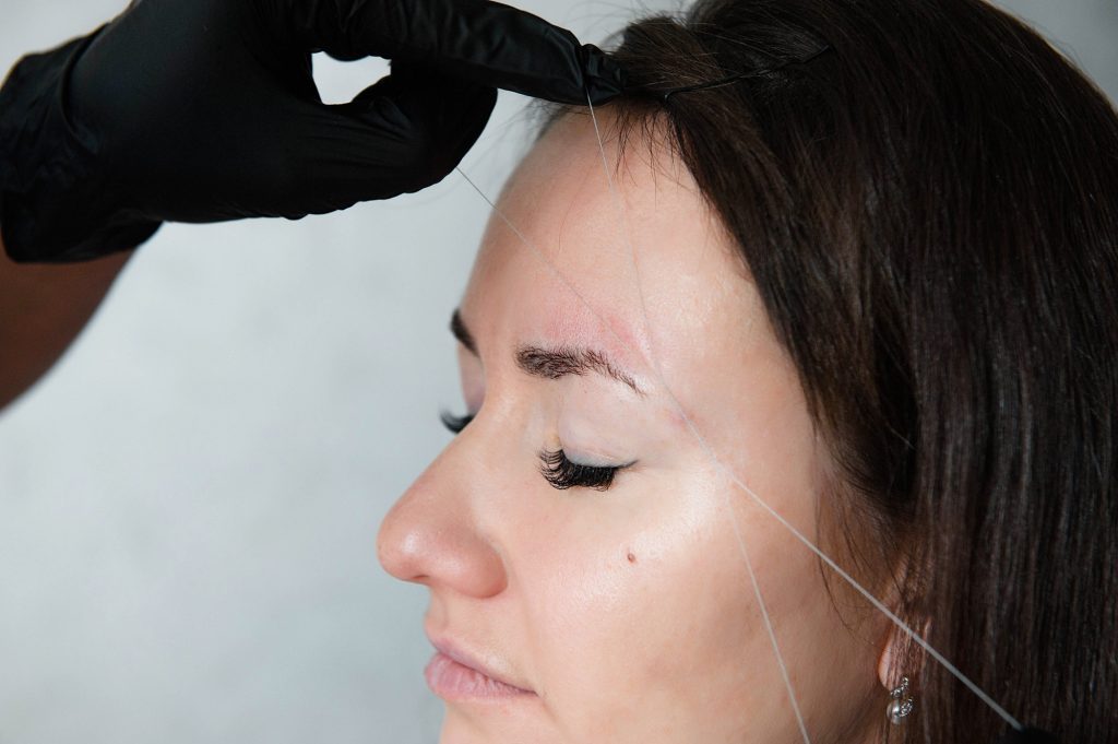 Browist makes eyebrow correction with tweezers thread. A model with dark hair in a light studio is s